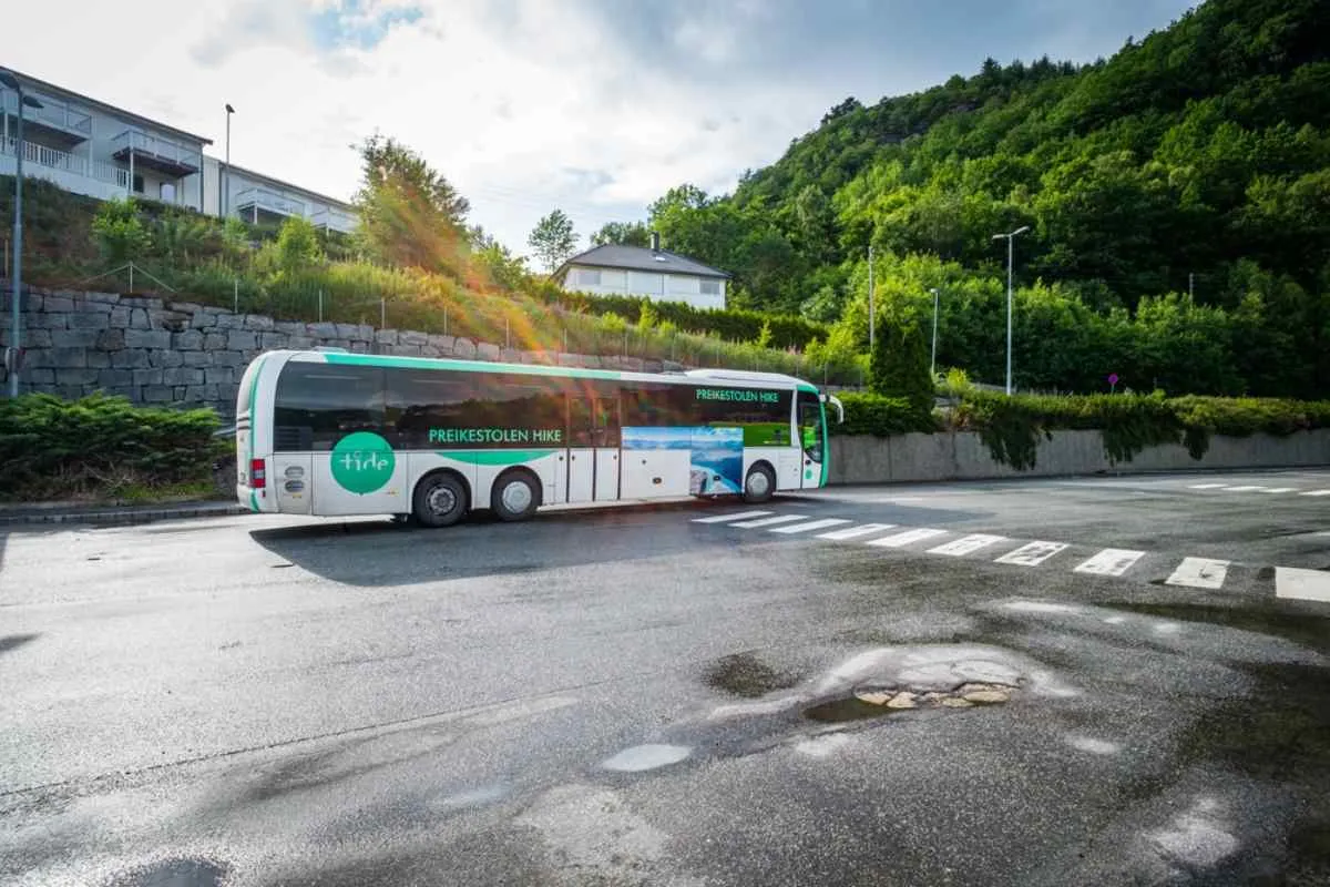 Bus departing toward the Preikestolen hike in Norway, parked along a quiet road with green hillsides and residential buildings nearby.