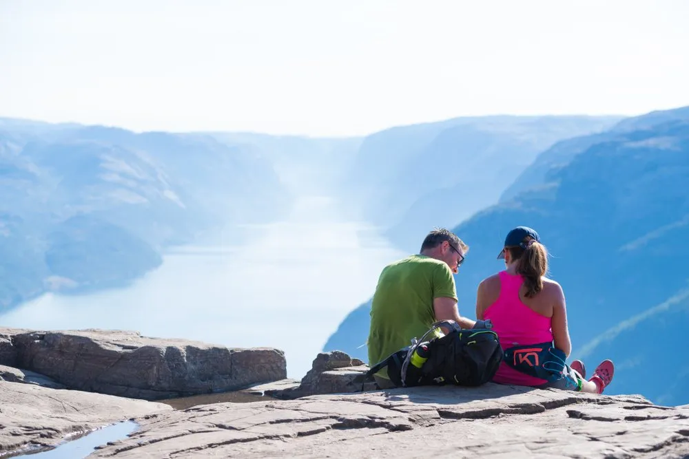 Couple sitting on the Preikestolen cliff, looking over Lysefjord on a clear day.