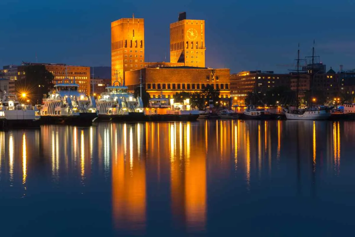Oslo City Hall at night with clock tower and lights reflecting on the harbor.