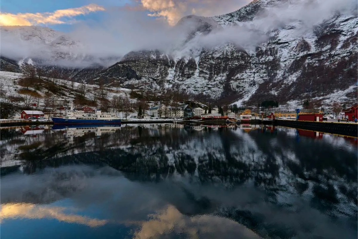 Small waterside village with red and white houses and a moored ferry, reflected in calm fjord water beneath snow-dusted peaks