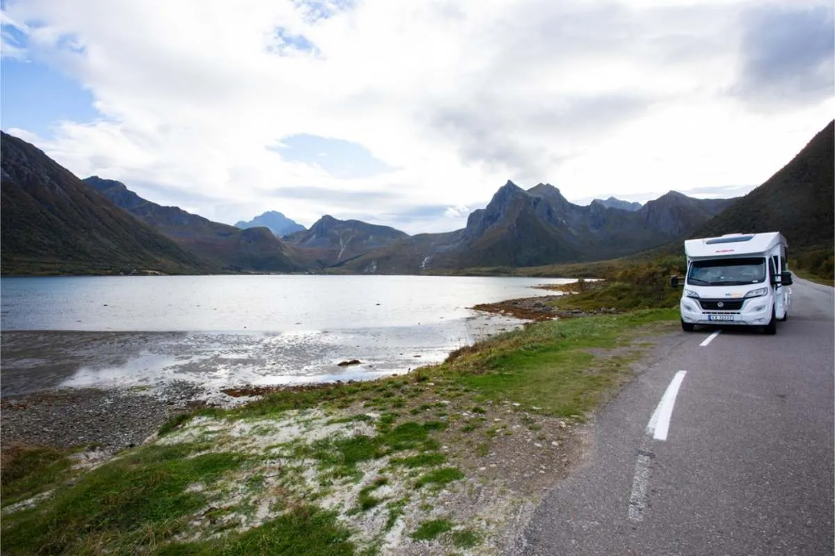 White motorhome on the roadside next to still fjord water, with jagged mountains along the far shore