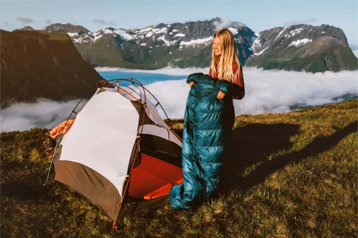 Woman unrolling a teal sleeping bag beside a small tent on a grassy ridge