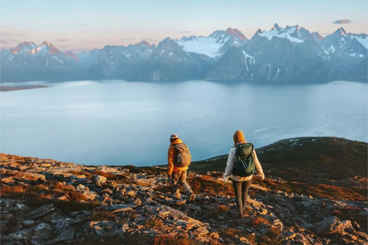 Two hikers with backpacks walking down a rocky slope at sunrise, overlooking a wide fjord and sharp snow-covered peaks