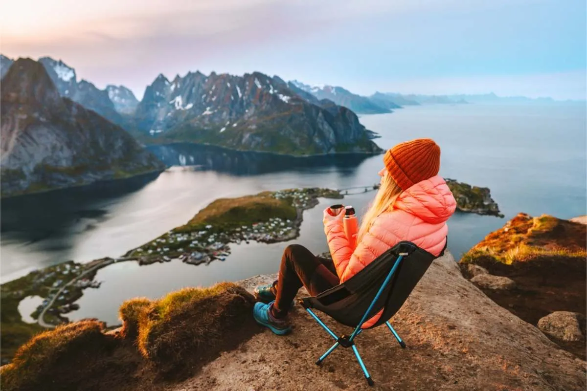 Woman in a pink jacket sitting in a folding chair on a clifftop with a thermos, looking down at a fishing village between fjords