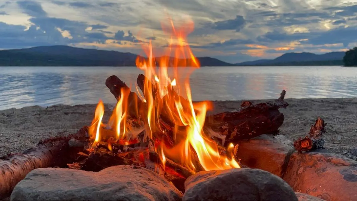 Open campfire burning between stones on a pebble beach, with a lake and dark hills behind under an evening sky