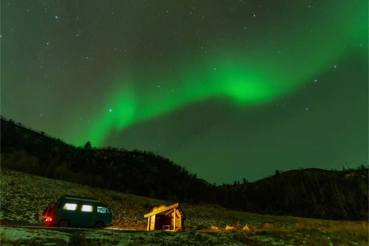 Green aurora across a starry sky over a van parked beside a small wooden shelter on a snow-patched hillside
