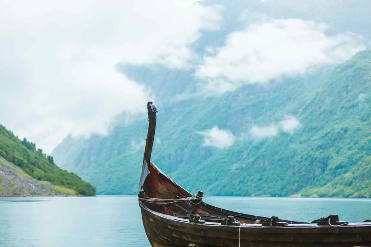 Traditional wooden Viking boat floating on a calm fjord, surrounded by steep green mountains and low clouds in Norway.