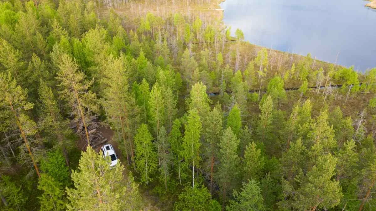Aerial view of a camper driving through a dense green forest near a lake, capturing the scale and remoteness of the Norwegian landscape.
