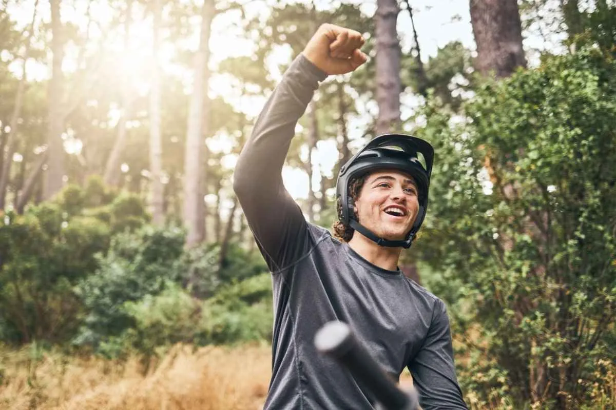 Smiling mountain biker in a helmet raising a fist on a forest trail, celebrating a ride outdoors.