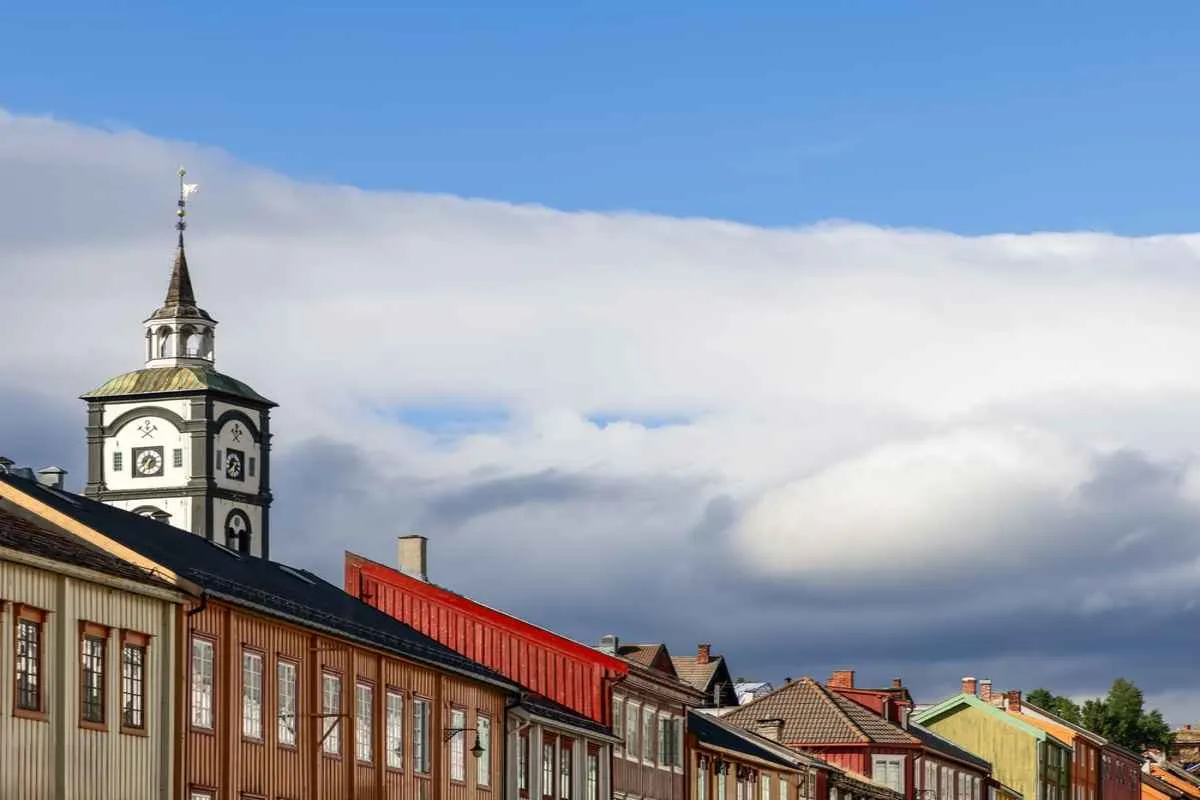 The Hyttklokka clock tower in Røros above a row of traditional wooden buildings under a blue sky with dramatic clouds.