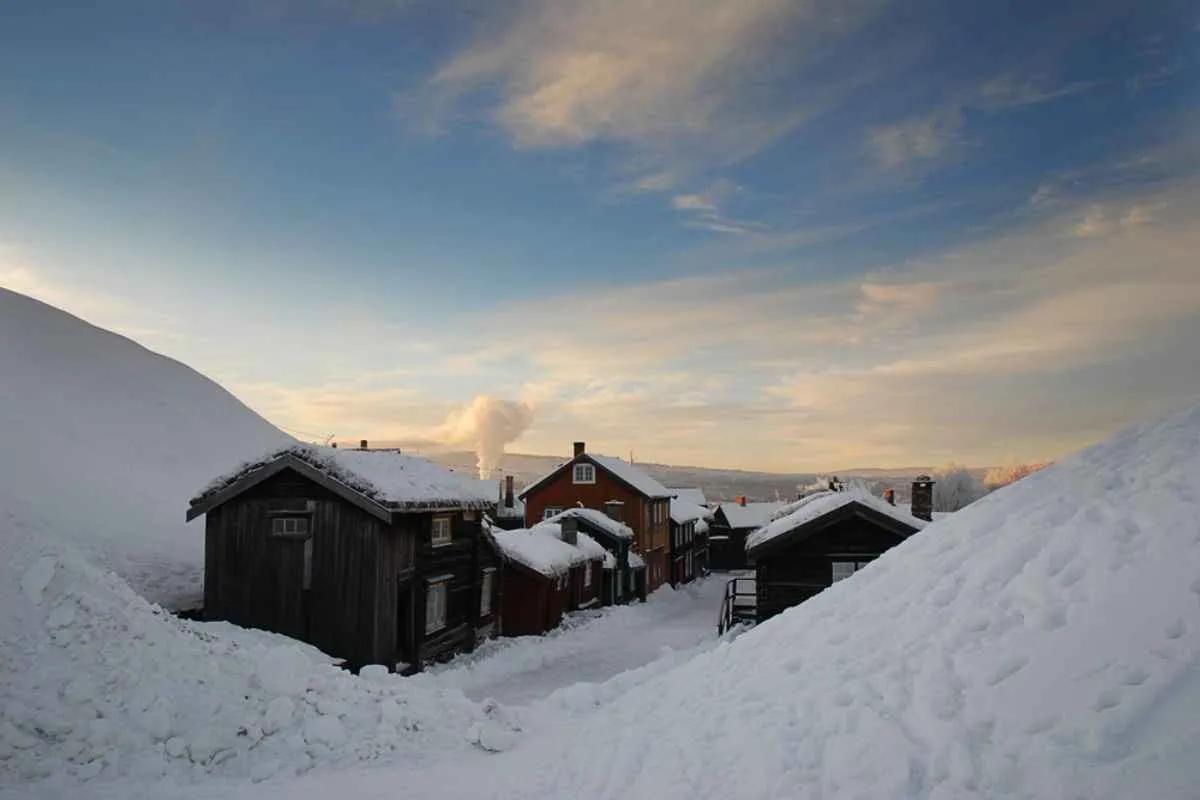 Snowy neighborhood street in Røros at sunset, with wooden houses buried in deep snow and smoke rising from chimneys.