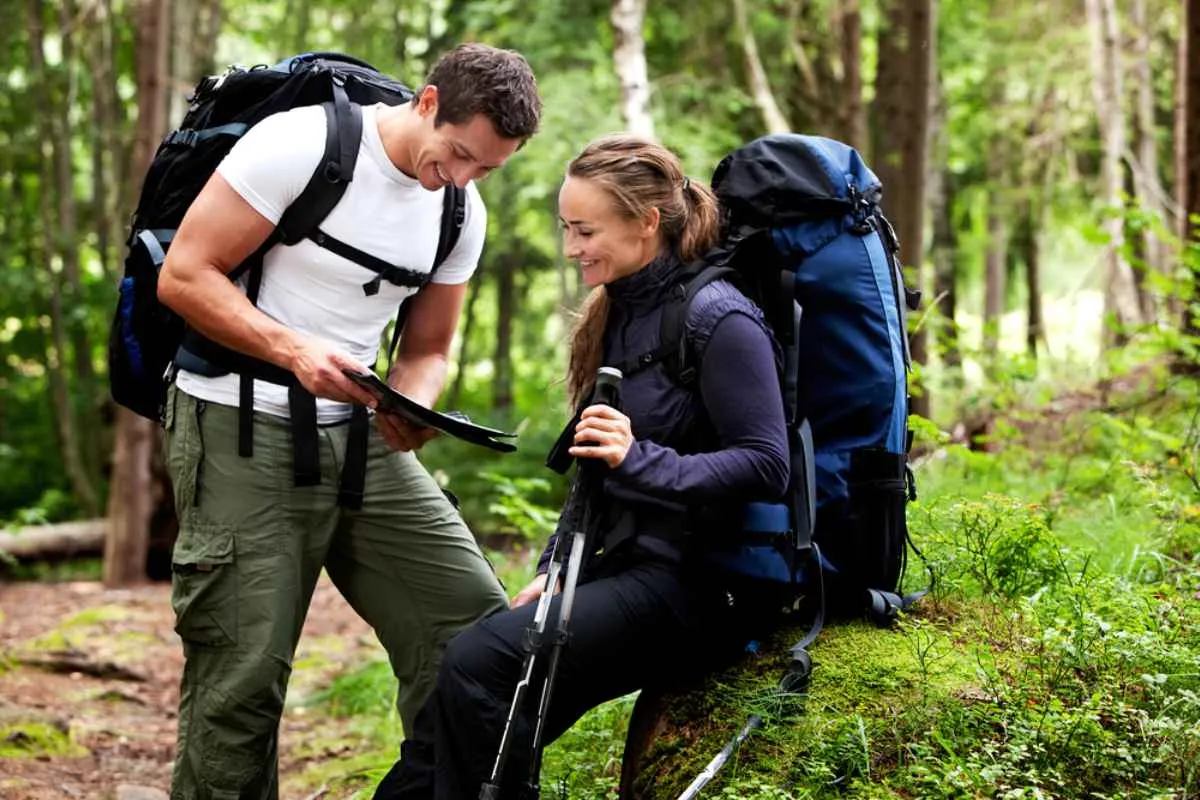 A hiking couple looks at a physical map in the forest while one of them rests on a rock.