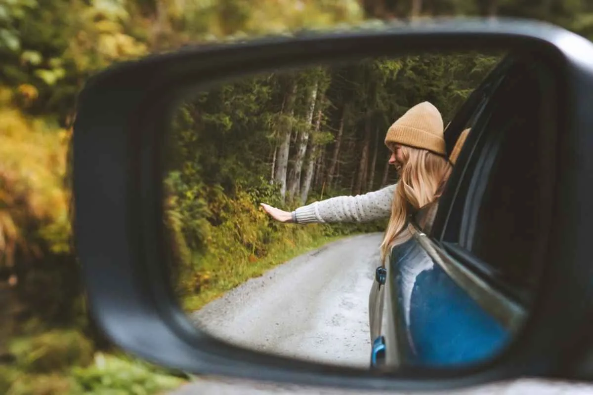 A blonde girl wearing a hat is reflected in the rearview mirror of a vehicle as she sticks her head out of the window.