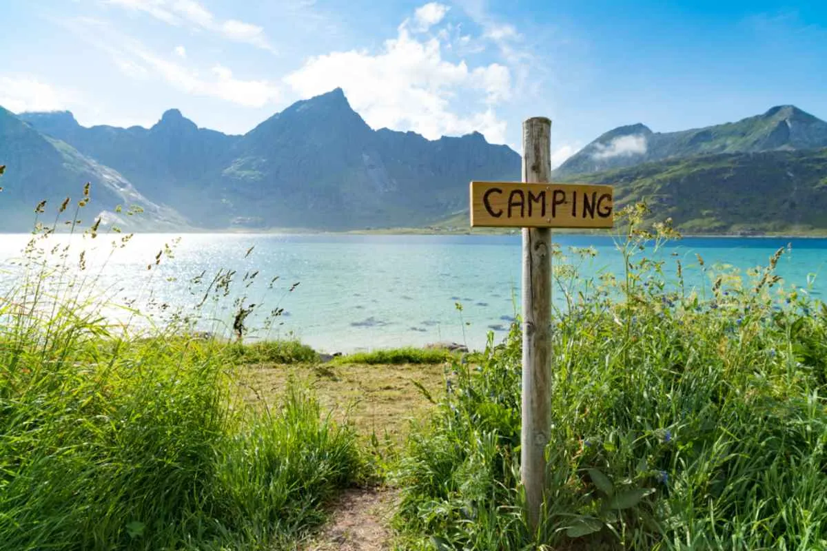 A sign on a Norwegian road indicates the possibility of camping in the area, surrounded by mountains, crystal-clear waters, and green vegetation.