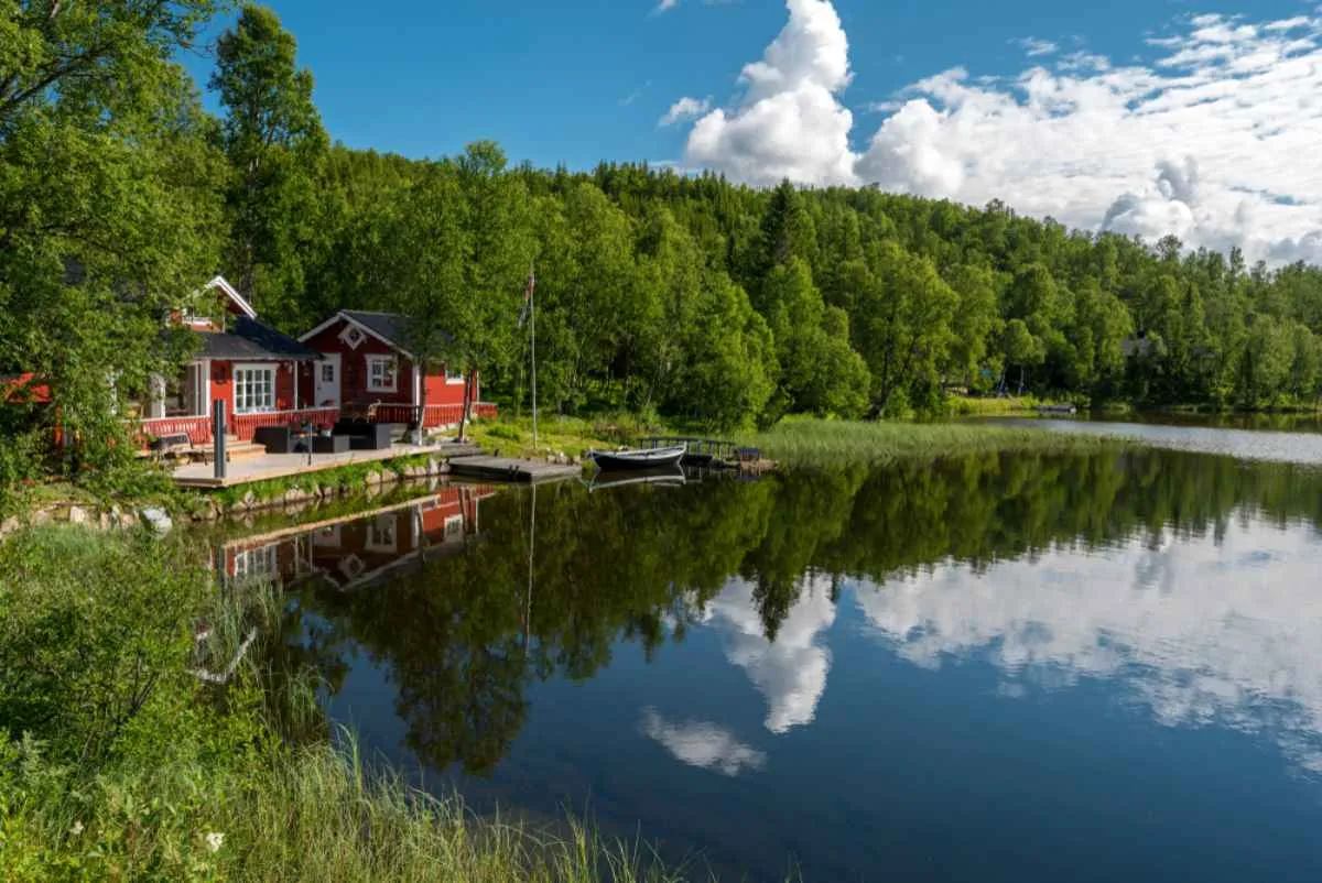 A typical Norwegian house in front of a calm lake that reflects both the clouds in the sky and the huge number of pines and other trees that surround it in nature.
