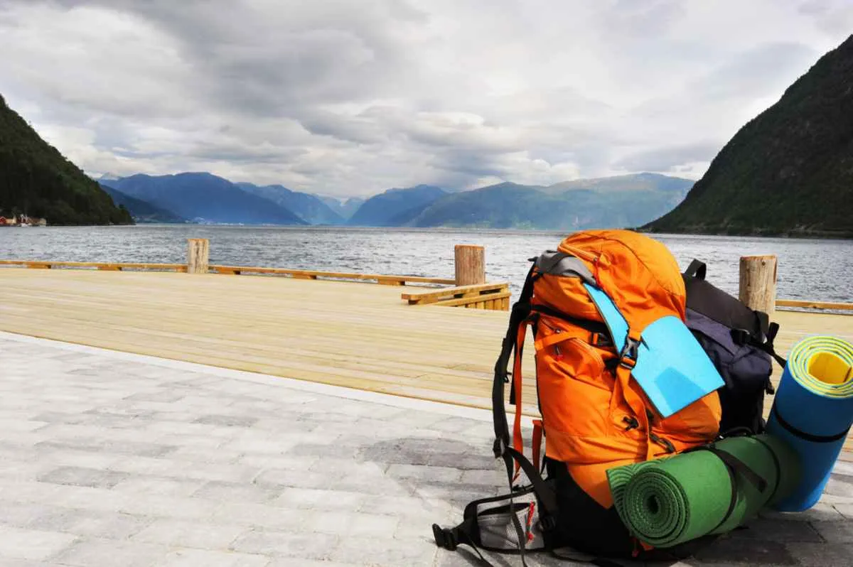 An orange hiker's backpack is positioned on the right side of the image near a Norwegian fjord on a sunny but cloudy day.