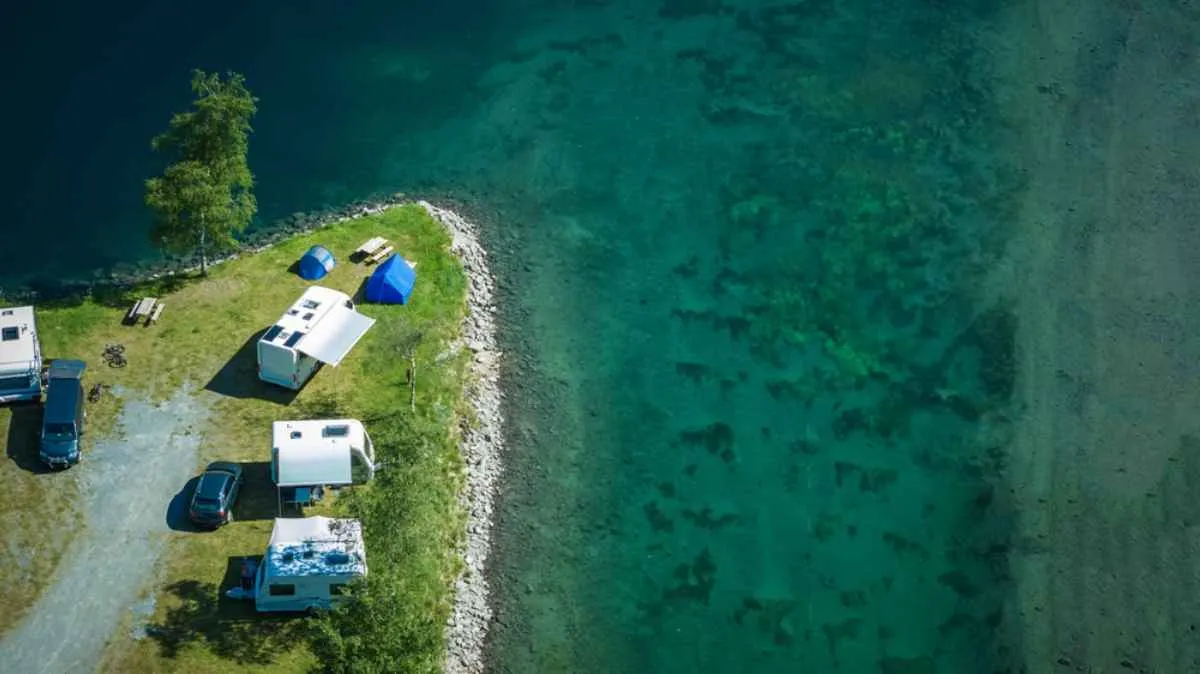 A group of white campervans sit on a plain located in front of a river of blue, sweet and calm waters to camp and enjoy the beautiful views that Norway has to offer.