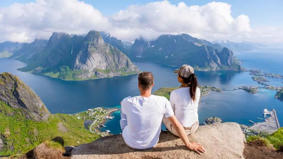 A tourist couple sits on what appears to be the top of a cliff as the Lofoten Islands of Norway rise above the turquoise waters of Norway.