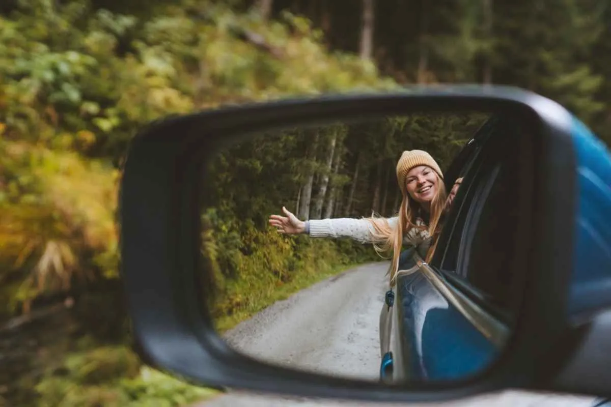 A blonde woman in a white cap and sweater peeks out part of her body through the window of a car as she sees herself reflected in the car's rearview mirror as they drive through a wooded road.