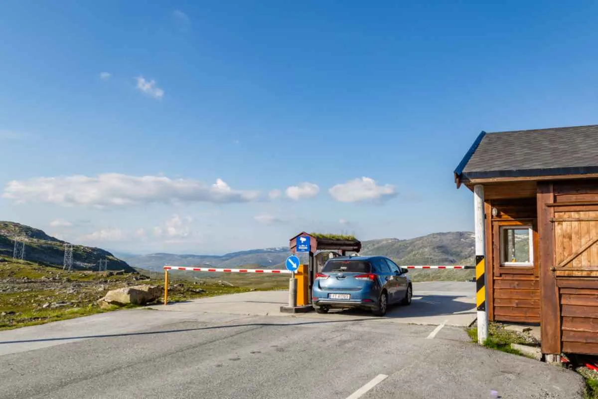 A blue vehicle remains parked in front of a toll booth on a secondary road in Norway in what looks like a rural area under a partly clear blue sky.