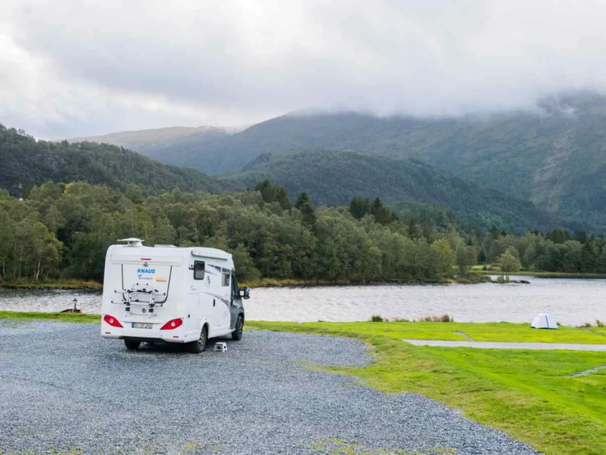 A white camper is parked in a Norwegian forest overlooking a lake that is itself surrounded by vegetation and mountains covered by low clouds.