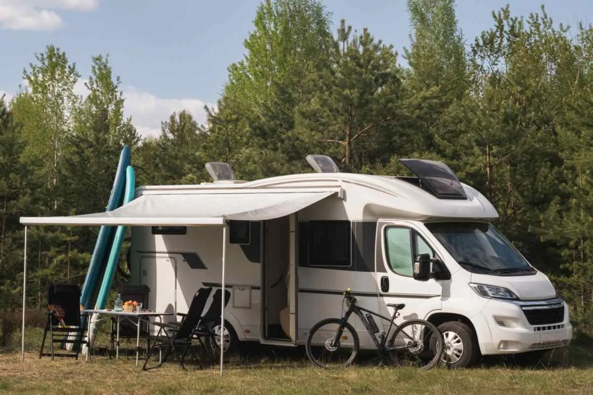 A motorhome parked in a Norwegian forest suitable for camping with girls and a table with fruit spread outside, as well as a bike on the right, surfboards on the left, and a large awning protecting its guests from the sun's rays in the center.