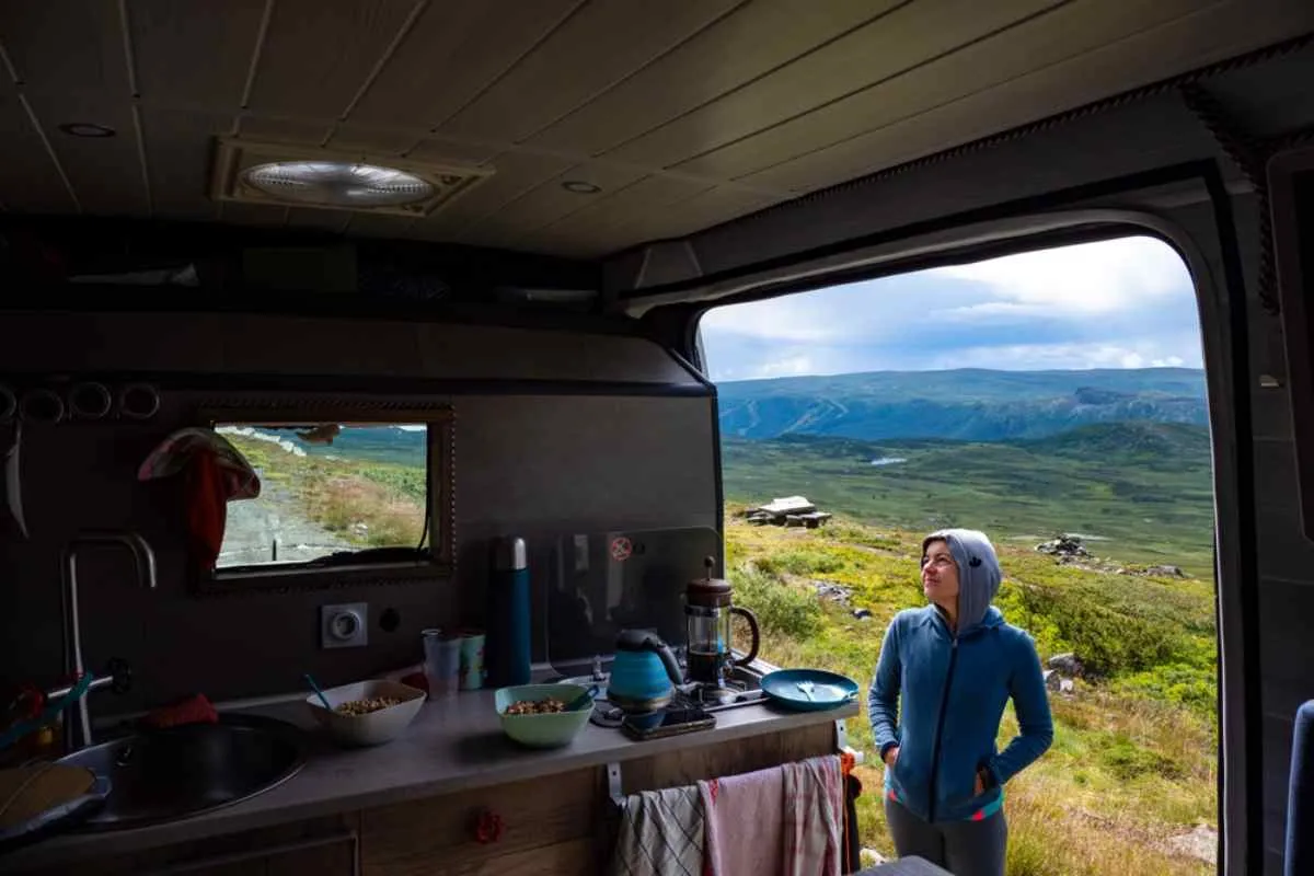 A hooded woman from the outside of her motorhome observes the interior of the motorhome, which features a well-equipped kitchen, as well as a window and door showing the beautiful Norwegian countryside around them.