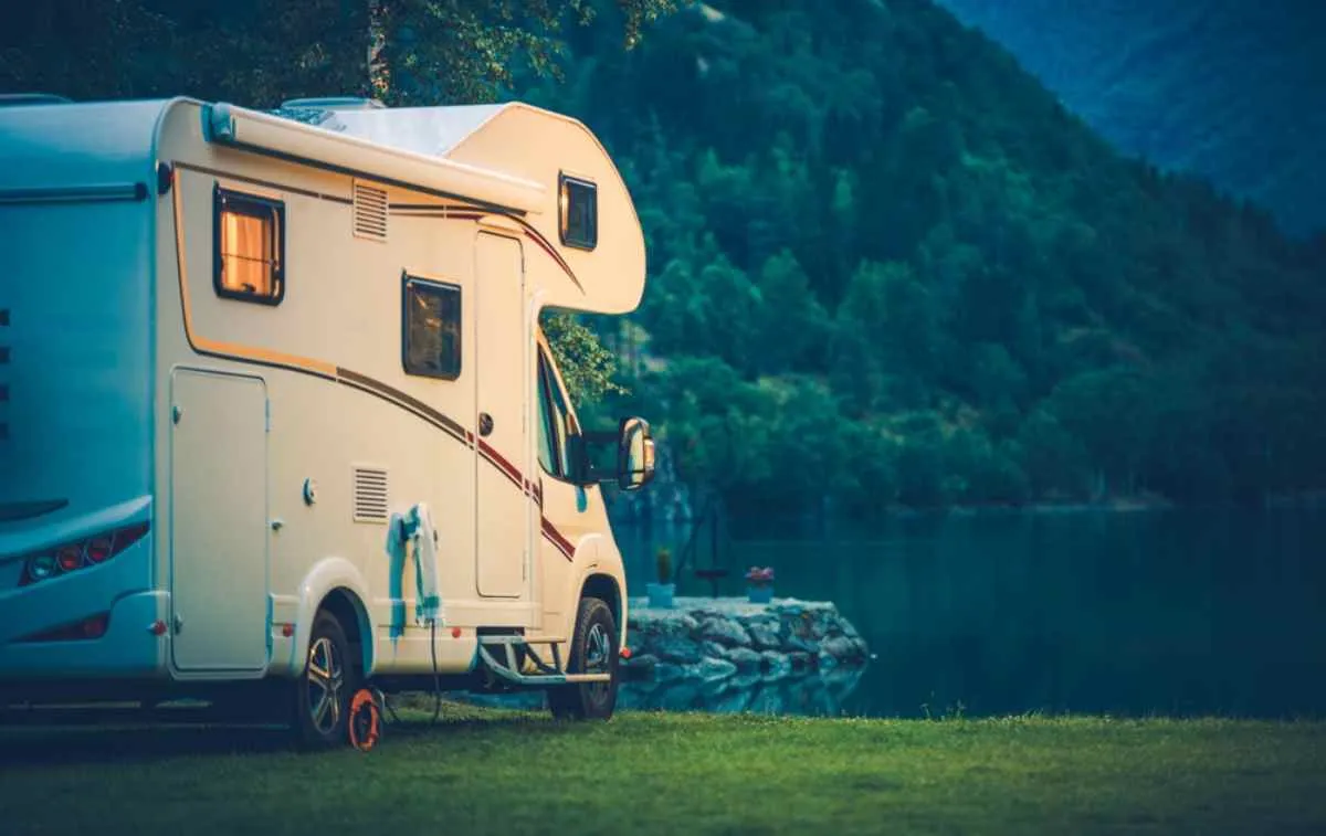 A white campervan is parked in the direction of a Norwegian lake in what looks like the last hours of the day, as the light is low and lights are on inside.