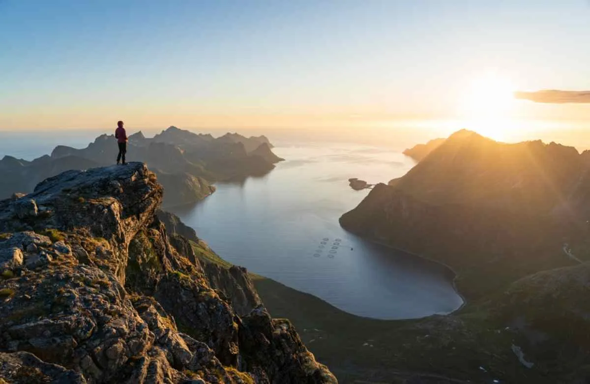 A hiker on top of a steep mountain observes the last rays of sunlight as they fall directly on the large body of water directly in front of him.