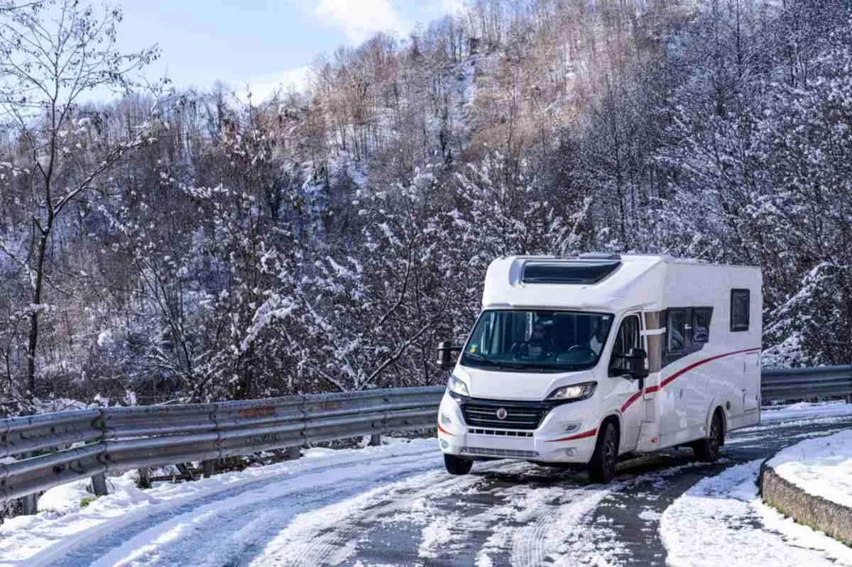 A white motorhome takes a curve on a completely snowy road near tall trees