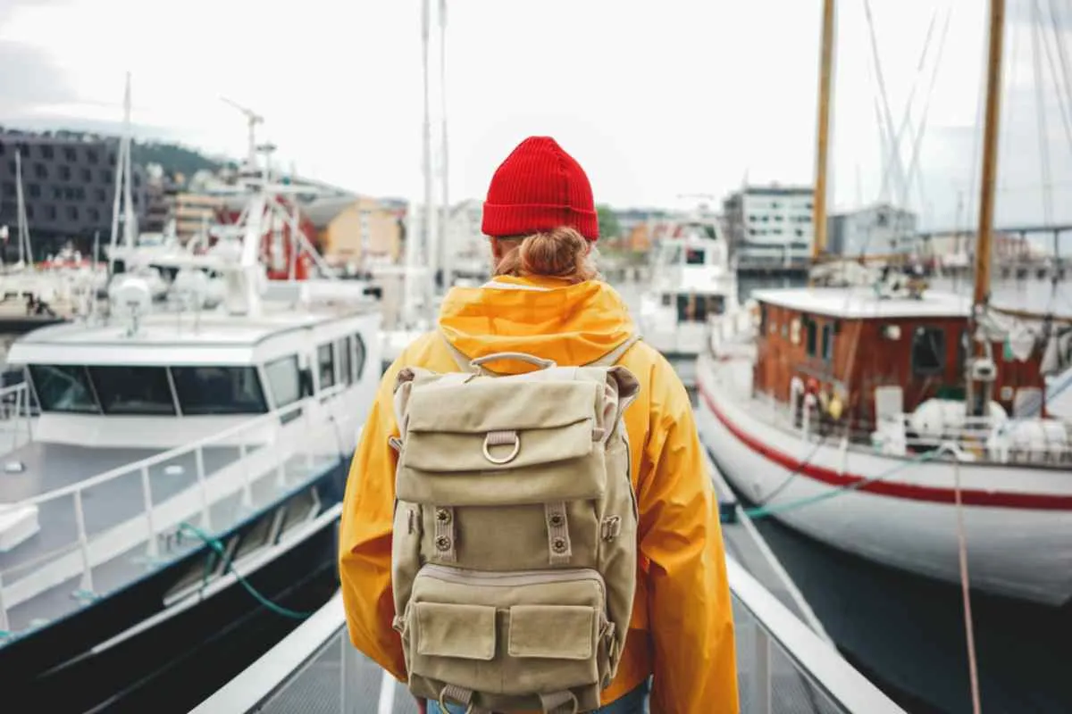 A person dressed in yellow, wearing a red cap and carrying a beige travel backpack walks straight ahead through what looks like a harbor with multiple ships around him