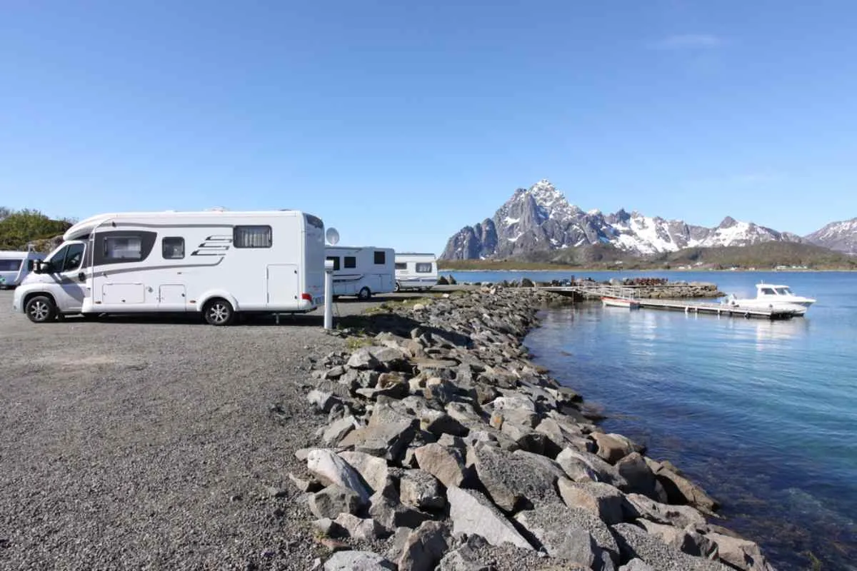 General view of a camper parked next to a beautiful lake with snow-capped mountains in the distance at Sandvika Fjord campsite.