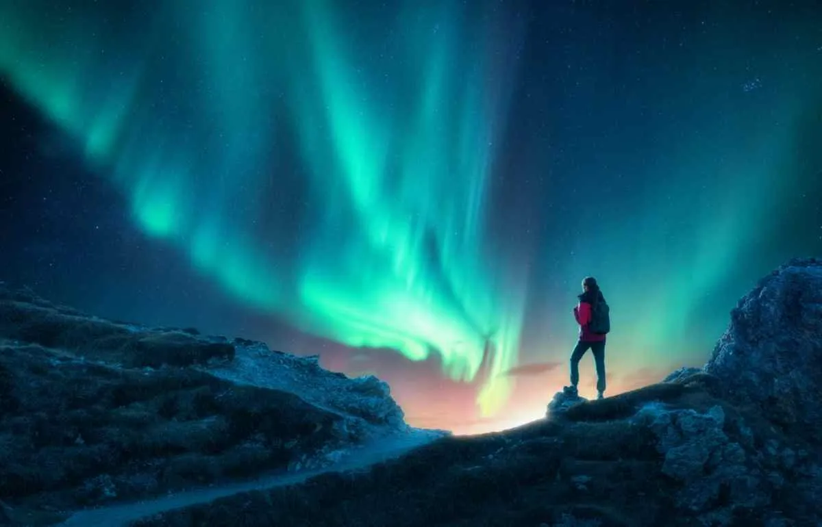 Close-up of what looks like a woman on top of a mountain observing the Norwegian Northern Lights in the middle of the night.