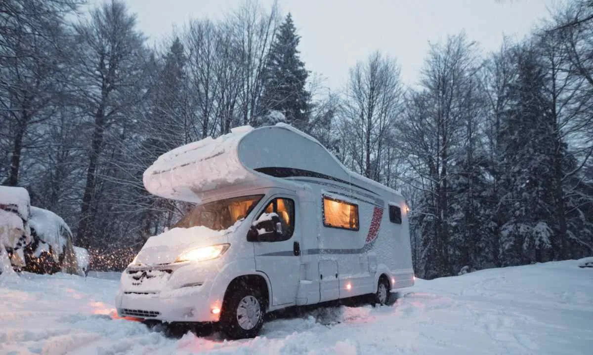 A white motorhome remains parked in a snowy Norwegian landscape with its headlights and interior lights on.