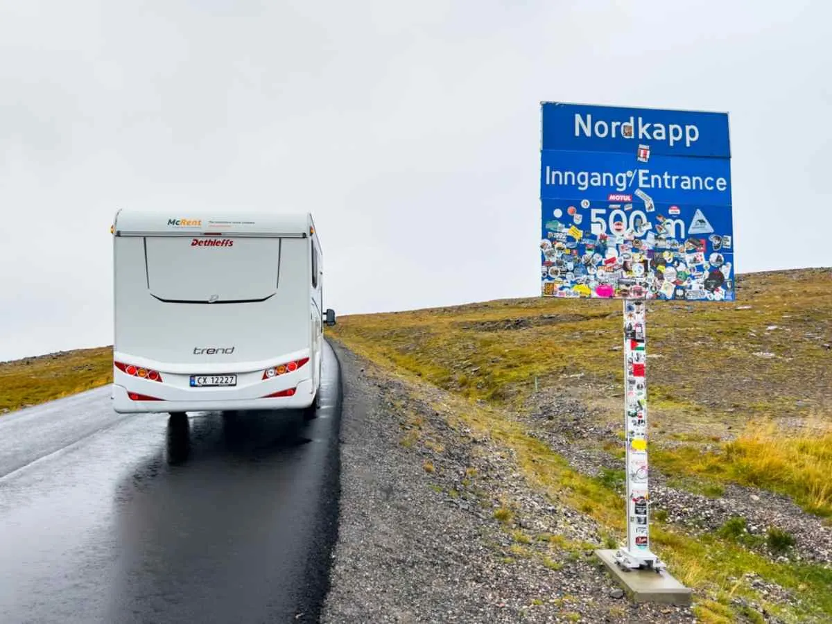 A white campervan crosses a stretch of Norwegian road with a sign announcing the arrival at Nordkapp.