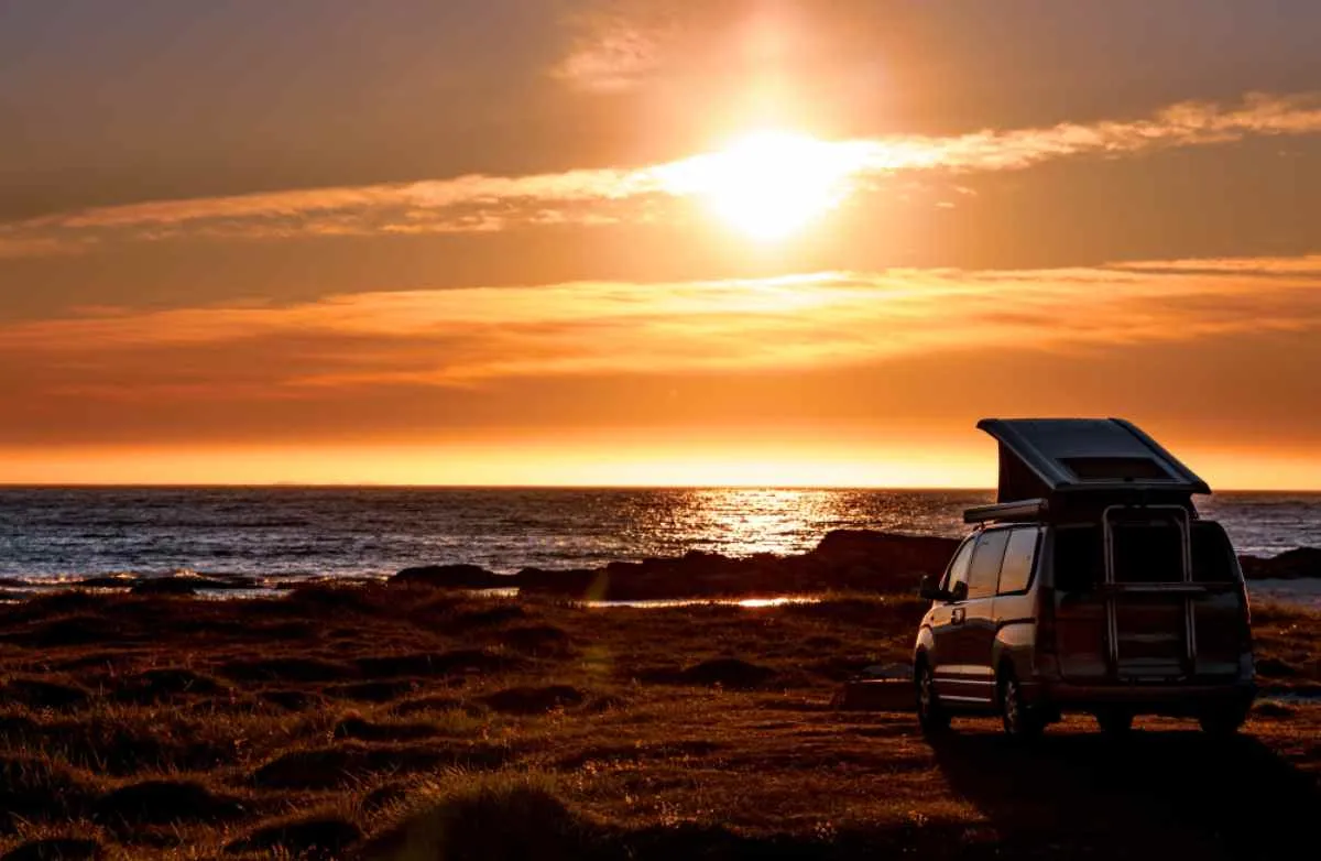 A vehicle with a roof tent remains parked on what looks like a cliff in front of a sunset over the sea.