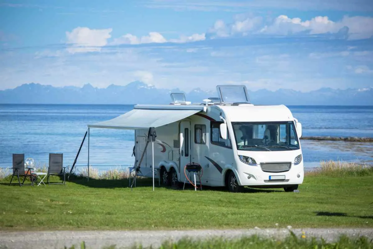 A white motorhome remains parked in a green Norwegian spot facing the open sea with camping tables and an awning spread over it.