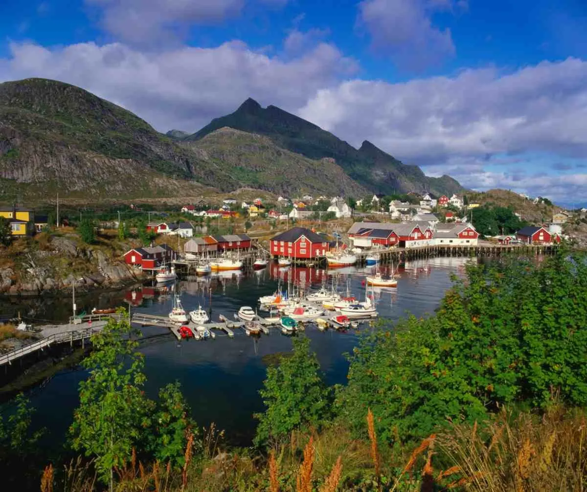 General view of the small Norwegian village of Sorvagen with a bay with docked boats and beautiful red wooden houses around it.