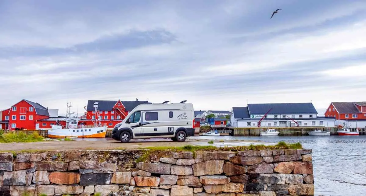 A compact campervan remains parked in a kind of harbor near a Norwegian village with reddish houses.