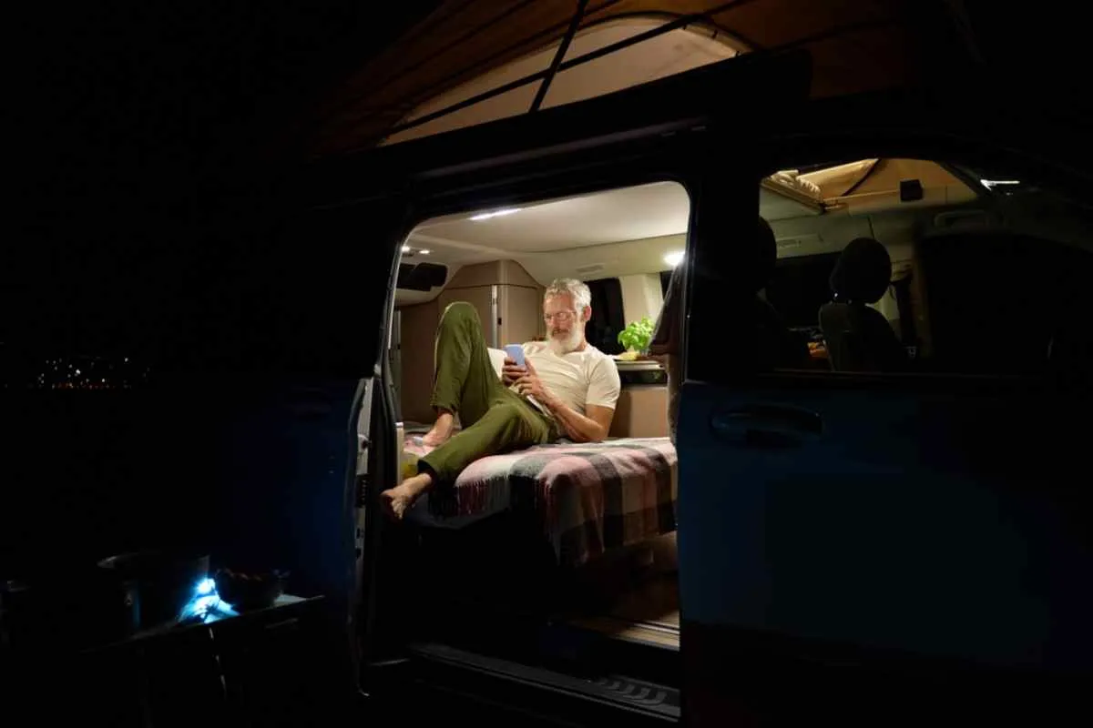 A bespectacled adult man stands inside his campervan lit up at night while staring at his cell phone half-lie down.
