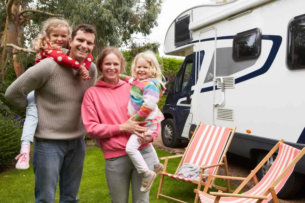 A family of two adults and two little blonde girls on their backs look on smiling as a motorhome parked with folding chairs is seen in the background.
