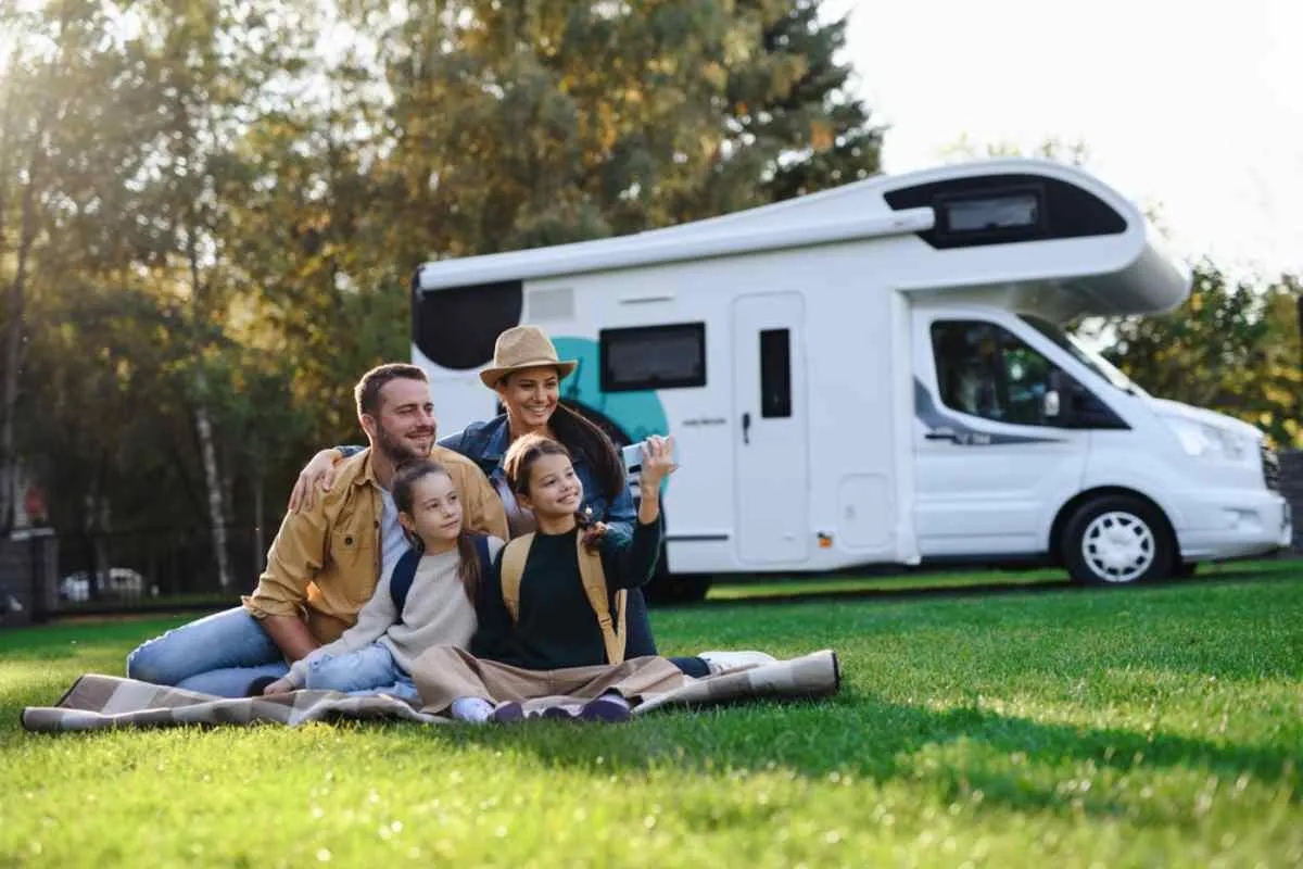 A family consisting of an adult man and woman with their young children sits on a lawn while the RV they are traveling in is parked next to them.
