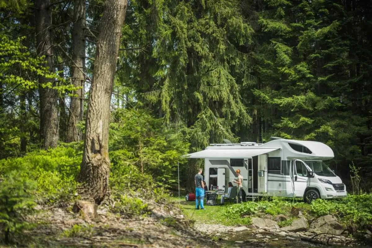 A motorhome remains parked in a green area that looks like a forest with huge trees surrounding it and a couple outside.