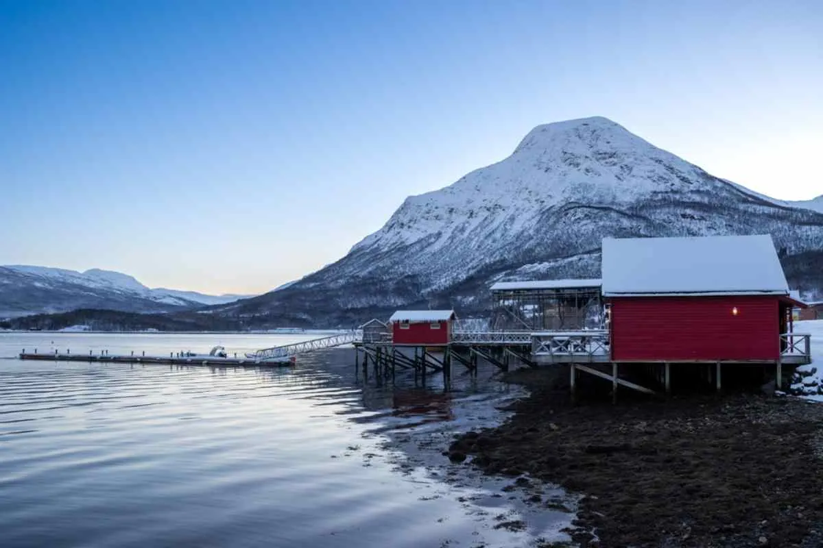 Overview of a completely snow-covered Norwegian landscape near a river with small red cottages on the bank.