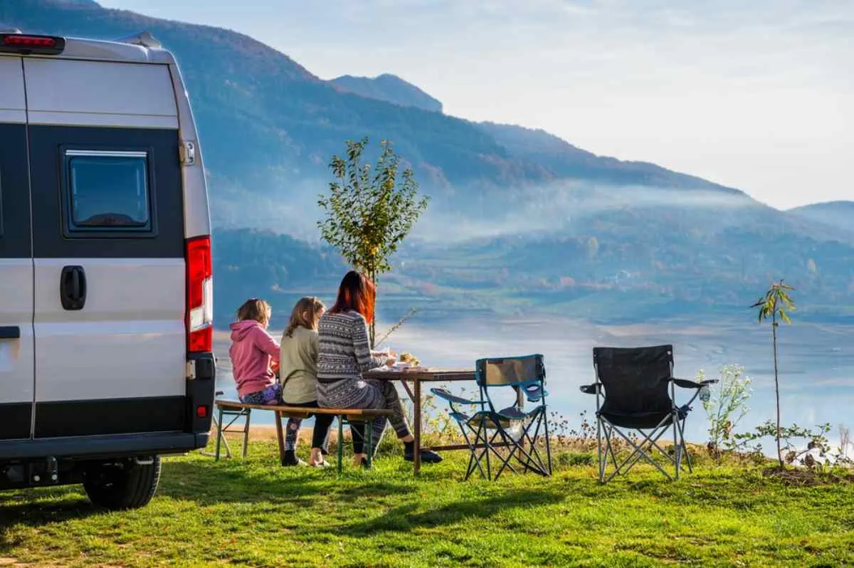 A family traveling in a camper van sits outside their parked vehicle, enjoying a campsite with folding chairs and tables in front of a river.