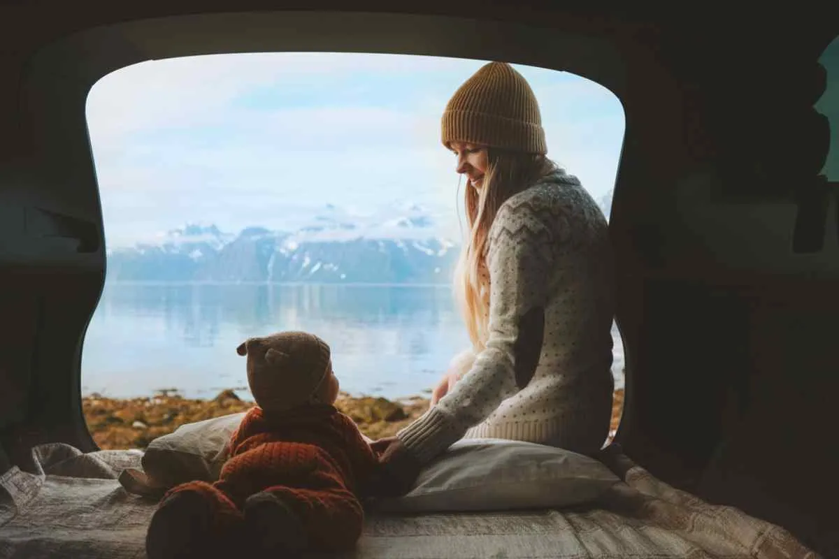 A mother and baby sit in the back of her camper van while a lake with snow-capped mountains can be seen in the distance.