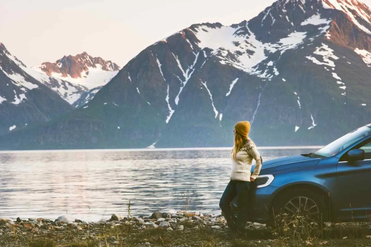 A woman stands outside her parked vehicle while a calm lake with mountains stands next to her.