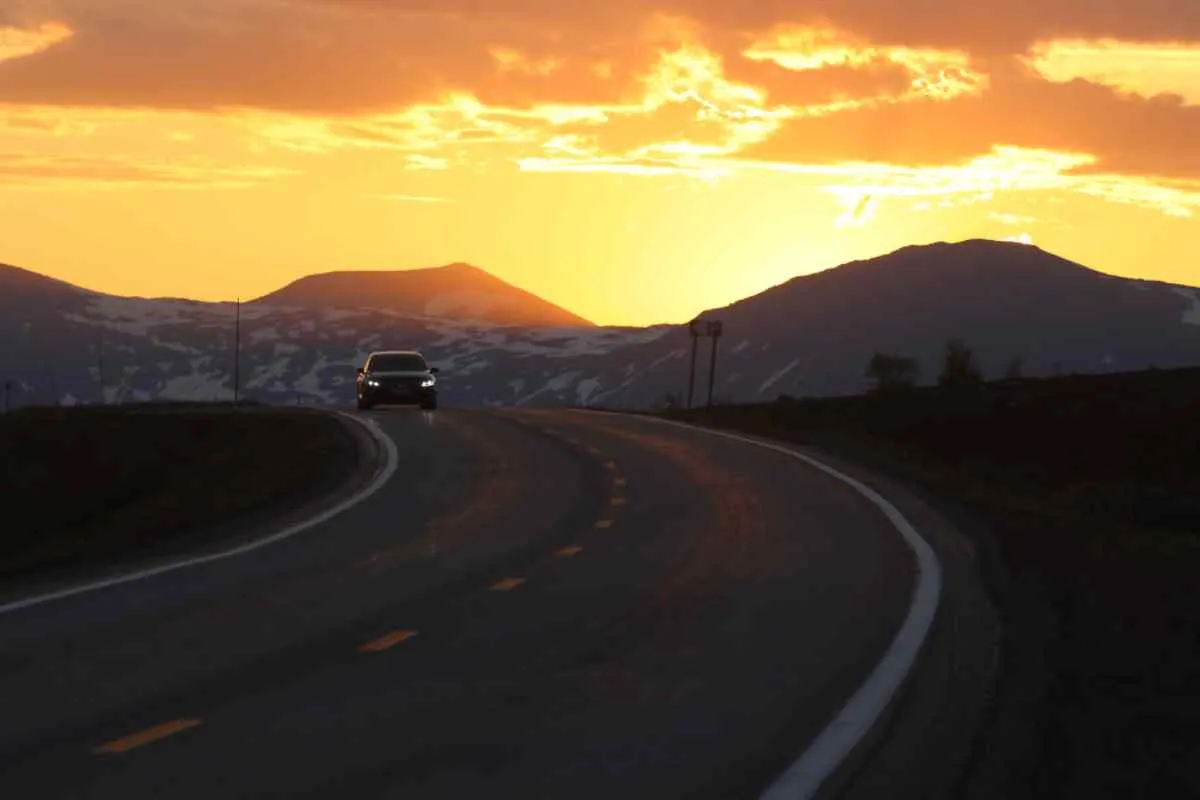 General view of the E6 road in Norway with the imposing midnight sun behind a car driving with lights on.