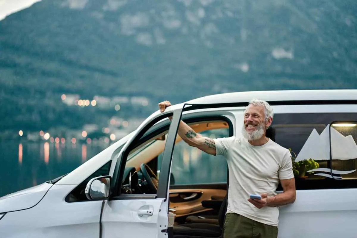 A gray-haired man smiles as he stands outside his parked white campervan with one hand on the open door and another on his cell phone.