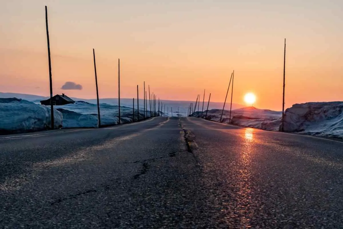 Shot from below of a Norwegian paved road with what looks like a snowy landscape on both sides and a sunrise in the background.